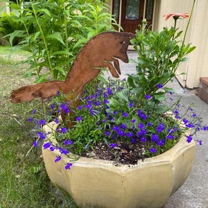 May include: A weathered metal fox sculpture leaps over a planter filled with vibrant purple flowers and green foliage. The hexagonal planter is a light yellow color, and a single pink flower blooms nearby. The scene is set outdoors.
