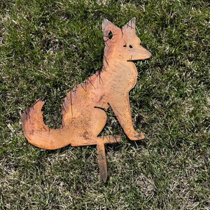 Puede incluir: Una estaca de jard&iacute;n de metal oxidado en forma de silueta de zorro. El zorro est&aacute; sentado con la cola enroscada alrededor de su cuerpo. La estaca est&aacute; insertada en el suelo.