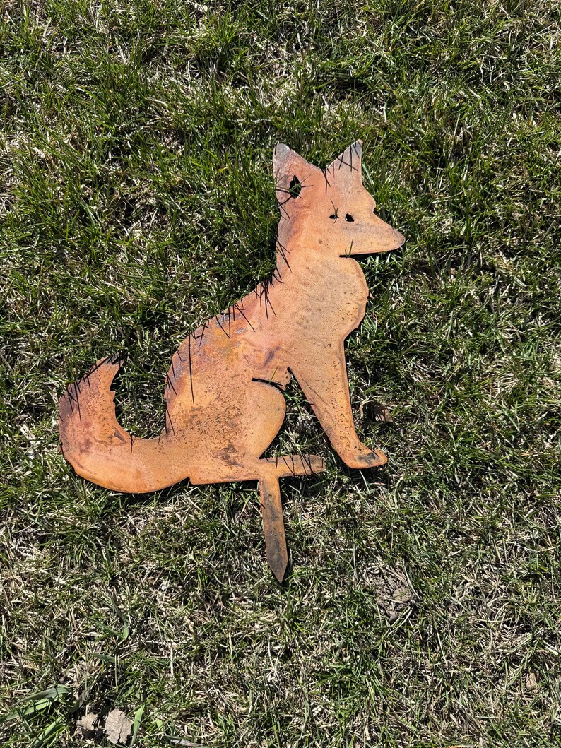 Puede incluir: Una estaca de jard&iacute;n de metal oxidado en forma de silueta de zorro. El zorro est&aacute; sentado con la cola enroscada alrededor de su cuerpo. La estaca est&aacute; insertada en el suelo.