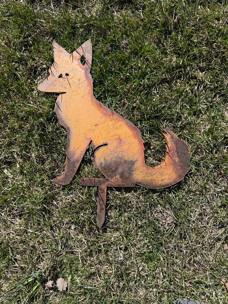 Puede incluir: Una estaca de jard&iacute;n de metal oxidado en forma de silueta de zorro. El zorro est&aacute; sentado con la cola enroscada alrededor de su cuerpo. La estaca est&aacute; insertada en el suelo.