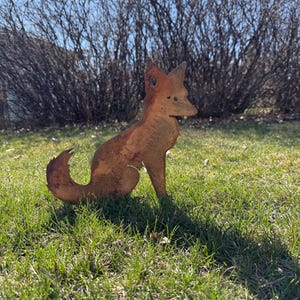Puede incluir: Una escultura de zorro de metal oxidado en posici&oacute;n sentada. El zorro est&aacute; mirando hacia la derecha y tiene una cola larga y tupida. La escultura est&aacute; sobre un lecho de hierba verde.