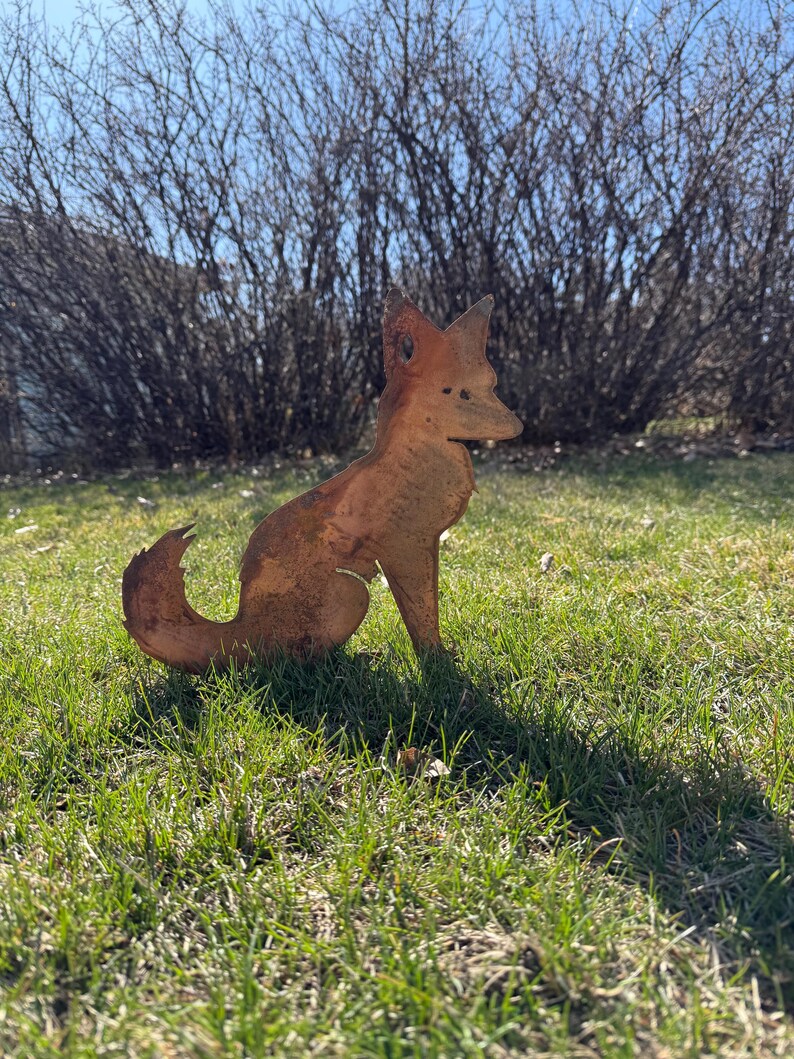 Puede incluir: Una escultura de zorro de metal oxidado en posici&oacute;n sentada. El zorro est&aacute; mirando hacia la derecha y tiene una cola larga y tupida. La escultura est&aacute; sobre un lecho de hierba verde.