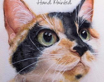 May include: A watercolor painting of a calico cat with black, orange, and white fur, green eyes, and long white whiskers. The text Hand Painted is written in black script above the cat's head. The cat is looking upward with a focused expression.
