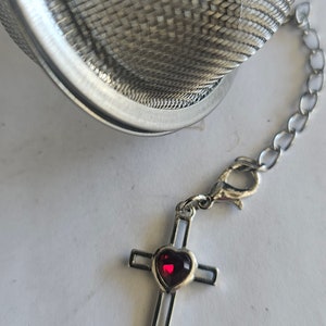 May include: A silver tea infuser with a chain and a silver cross charm with a red heart-shaped gem.