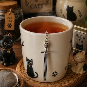 Peut inclure: Une tasse en céramique blanche remplie de thé ambré, ornée d'illustrations de chats noirs et d'empreintes de pattes. Un infuseur à thé en métal argenté en forme d'épée avec un pommeau tête de chat pend au bord, relié par une chaîne à une breloque patte.