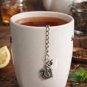 May include: A white ceramic mug filled with tea, featuring a silver dragon tea infuser. A slice of lemon, a jar of honey, and a teapot are in the background. Tea leaves and mint leaves are also visible.