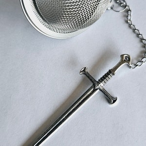 May include: A silver-colored metal tea infuser featuring a spherical mesh basket and a long, thin chain attached to a detailed sword-shaped charm. The sword charm has a crossguard and a textured handle, all finished in a polished metallic silver tone.