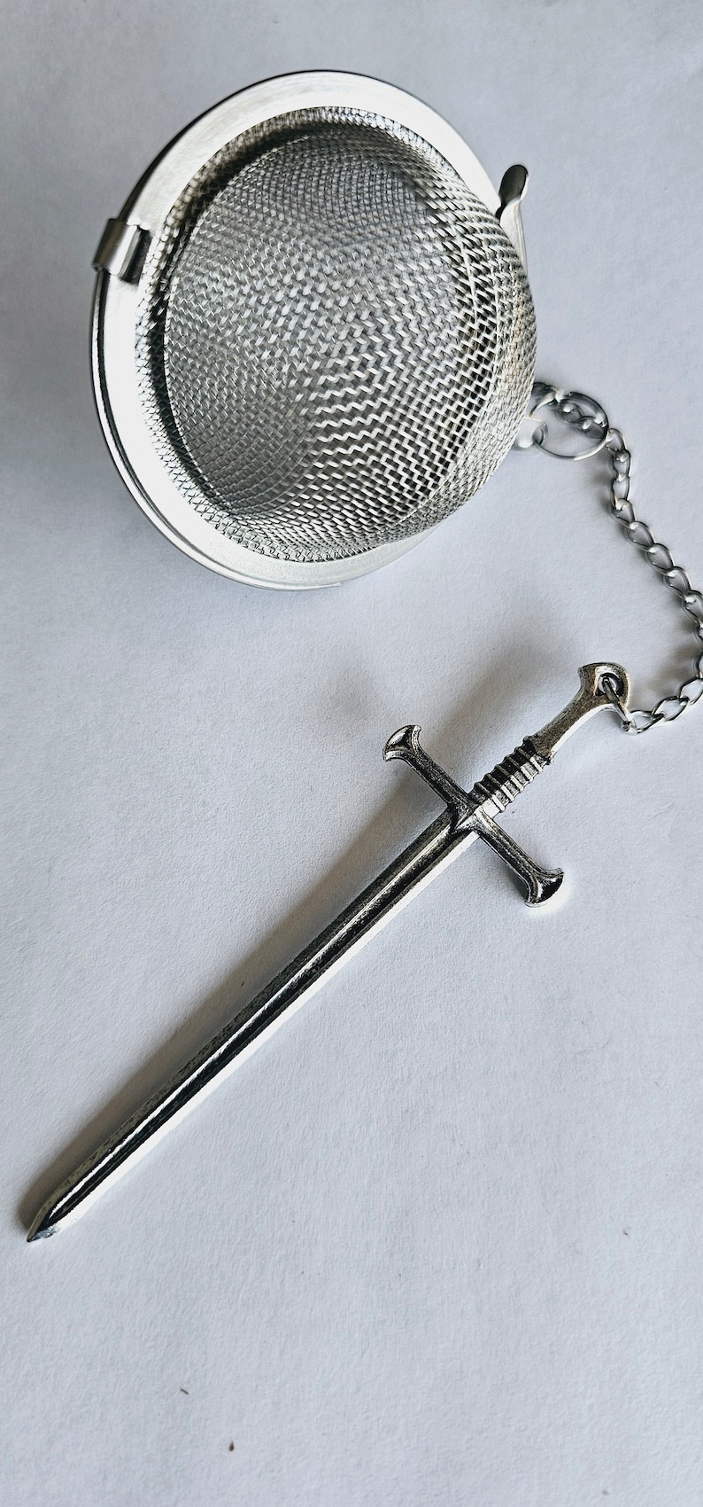 May include: A silver-colored metal tea infuser featuring a spherical mesh basket and a long, thin chain attached to a detailed sword-shaped charm. The sword charm has a crossguard and a textured handle, all finished in a polished metallic silver tone.