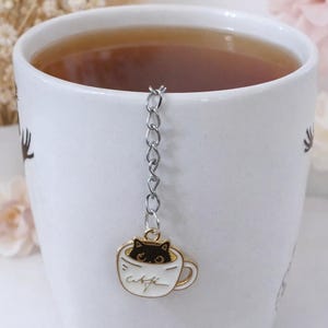 以下が含まれることがあります： お茶が入った白いマグカップ。カップの中の猫のティーインフューザーが付いています。インフューザーは、小さな金色の縁取りのカップで、中に黒い猫が入っており、銀色のチェーンで吊り下げられています。「cafe」という文字がカップに書かれています。