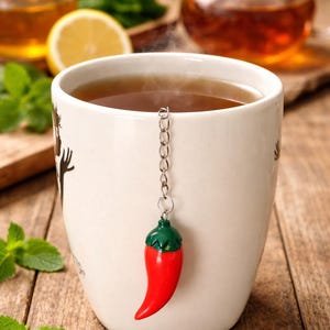 Könnte beinhalten: Eine weiße Keramiktasse gefüllt mit Tee, mit einem roten Chili-Pfeffer-Tee-Ei. Das Bild zeigt eine gläserne Teekanne, Honig, Zitrone und Minzblätter, alles auf einer Holzoberfläche. Das Tee-Ei ist an einer Kette befestigt.