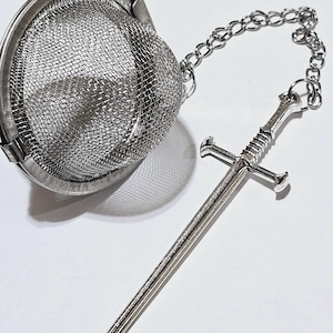 May include: A silver-toned metal tea infuser featuring a spherical mesh basket with a hinged lid. A silver metal chain is attached to the infuser, ending in a detailed miniature sword charm with a crossguard and a textured handle grip.
