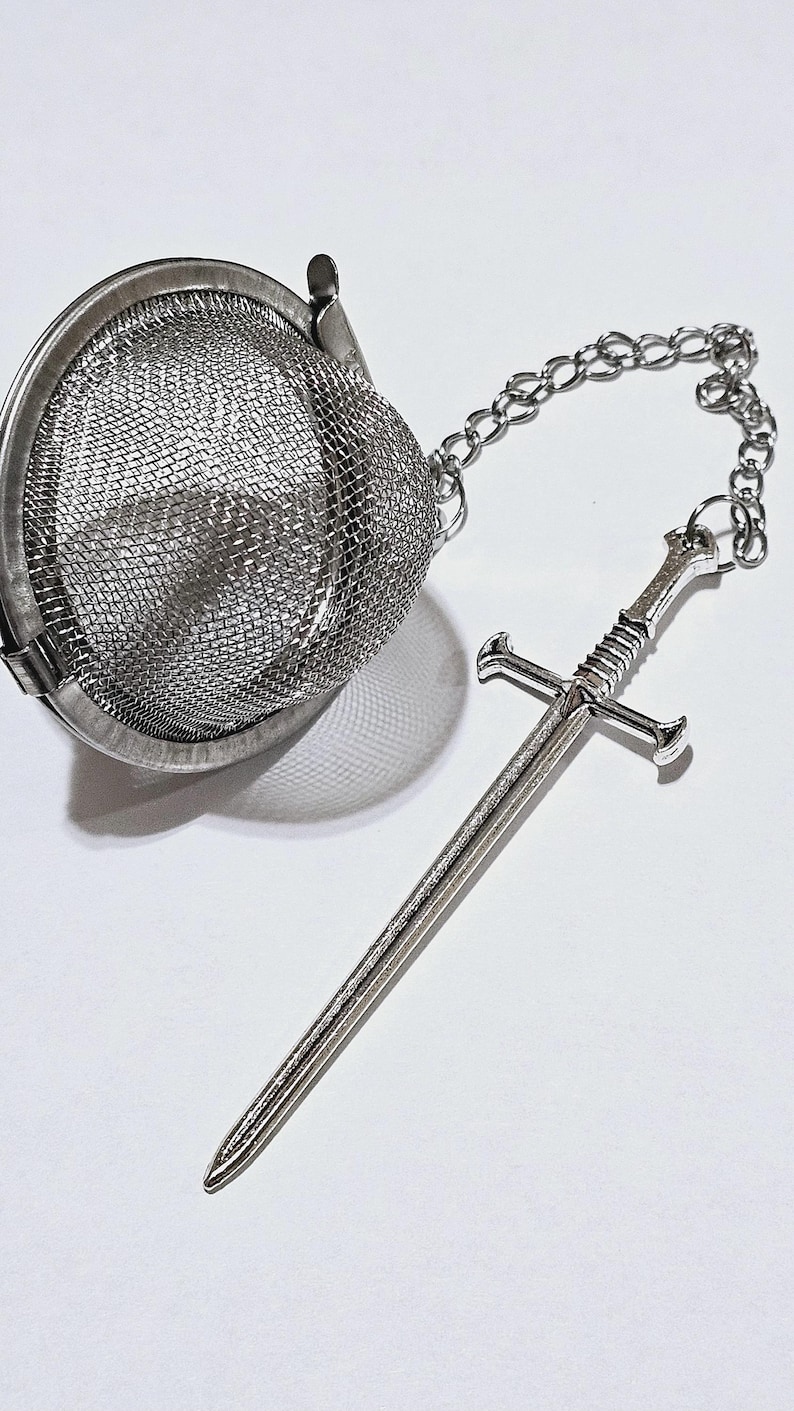 May include: A silver-toned metal tea infuser featuring a spherical mesh basket with a hinged lid. A silver metal chain is attached to the infuser, ending in a detailed miniature sword charm with a crossguard and a textured handle grip.