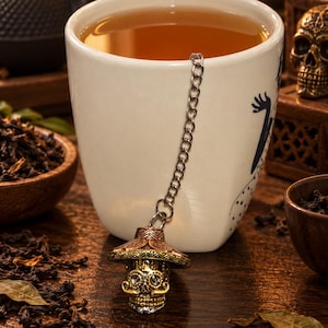 May include: A metal tea infuser shaped like a skull wearing a sombrero, attached to a silver-toned chain. The skull features intricate engraved details and a mustache, with a wide-brimmed hat resting on top. The infuser hangs over the edge of a white ceramic mug filled with amber-colored tea.