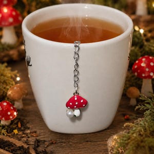 Peut inclure: Une tasse en céramique blanche remplie de thé fumant, avec une chaîne en métal argenté suspendue sur le bord. Attaché à la chaîne se trouve un petit pendentif champignon rouge à pois blancs avec une perle blanche suspendue en bas.