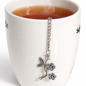 May include: A white ceramic mug filled with tea, with a silver floral tea infuser hanging inside. Steam rises from the tea. The mug has black decorative elements on the side.