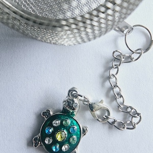 May include: A silver tea infuser with a chain and a turtle charm. The turtle is green with a blue and yellow gem design.