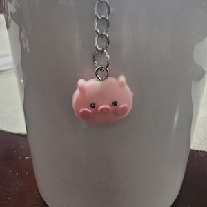 Könnte beinhalten: Eine weiße Keramiktasse mit einem rosa Schweinchen-Anhänger, der am Rand hängt. Der Schweinchen-Anhänger ist an einer silbernen Kette befestigt.