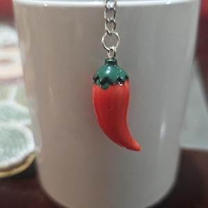 Könnte beinhalten: Ein roter Chili-Pfeffer-Tee-Infuser mit grünem Stiel, der an einer silbernen Kette hängt, hängt über einer weißen Tasse. Der Chili-Pfeffer hat eine leuchtend rote Farbe.