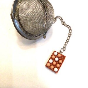 May include: A silver tea infuser with a chain attached to a brown egg carton charm. The egg carton charm has white and brown eggs inside.