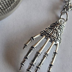 Puede incluir: Infusor de té de plata con un diseño de mano de esqueleto unido a una cadena. El infusor es redondo y tiene un fondo de malla.