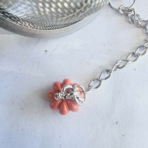May include: A silver tea infuser with a chain and a small, orange, flower-shaped charm. The infuser is made of fine mesh and has a round shape.