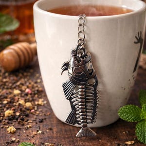 May include: A white ceramic mug filled with tea, with a decorative silver fish-shaped tea infuser hanging inside. The mug sits on a wooden surface scattered with tea leaves and herbs. Steam rises from the tea.