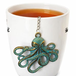 May include: A white ceramic mug filled with tea, featuring a decorative octopus tea infuser. The octopus infuser is bronze and teal, attached to a chain. The mug has black decorative accents.