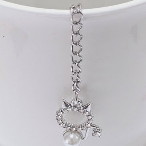 Puede incluir: Un charm de plata con una perla y pedrería en forma de gato. El charm cuelga de una cadena de plata.