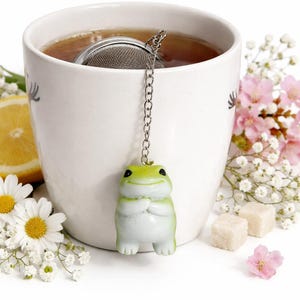 May include: A white ceramic mug filled with tea, featuring a stainless steel tea infuser with a green frog charm. The scene includes daisies, lemon slices, pink flowers, and sugar cubes, creating a serene tea-time setting.