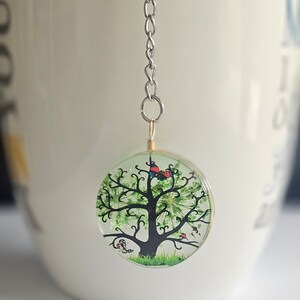 Könnte beinhalten: Eine silberne Kette mit einem runden Glasanhänger, der einen handgemalten Baum mit grünen Blättern, einem braunen Stamm und roten und blauen Vögeln zeigt.