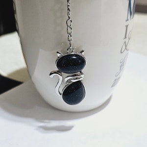 Puede incluir: Un infusor de té de plata con forma de gato con un cuerpo de piedra preciosa negra que cuelga de una cadena de plata en una taza de té blanca.
