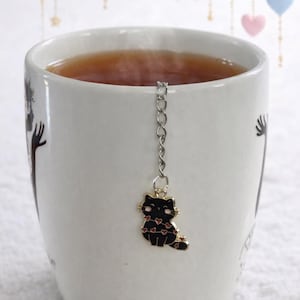May include: A white ceramic mug filled with tea, with a black cat tea infuser decorated with red hearts. The infuser hangs from a silver chain. Decorative stars and hearts are in the background.