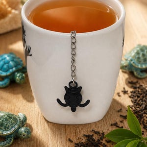 Peut inclure: Une tasse en céramique blanche remplie de thé ambré, dotée d'une chaînette en métal argenté attachée à une breloque infuseur à thé en forme de tortue noire suspendue au bord. La tasse est décorée de petites silhouettes de tortues noires.