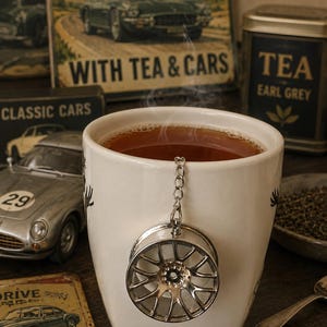 May include: A white ceramic mug filled with steaming dark tea, featuring a silver-colored metal tea infuser shaped like a car wheel rim hanging from a chain over the front rim of the mug, with a visible center hole and multiple spokes.