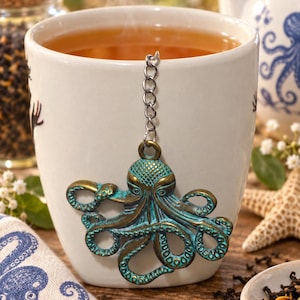 May include: A white ceramic mug filled with steaming tea, featuring a metal tea infuser shaped like an octopus. The octopus charm has a weathered bronze and teal patina finish and is attached to a silver-toned metal chain hanging over the rim.