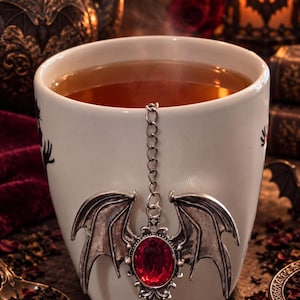 Puede incluir: Una taza de cerámica blanca llena de líquido ámbar, con un accesorio de infusor de té de metal plateado. El infusor tiene una piedra preciosa roja ovalada central rodeada de alas de murciélago plateadas y una cadena de metal.