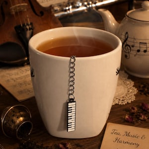 May include: A white mug of tea with a piano key tea infuser. A teapot, violin, and sheet music are in the background. A card reads "Tea, Music & Harmony". The image has a warm, musical theme, with a focus on tea and music.