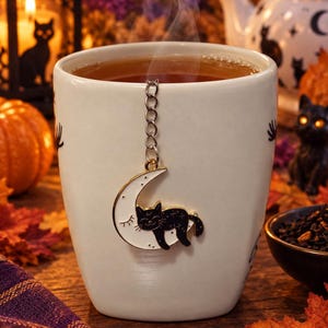 Puede incluir: Una taza de cerámica blanca llena de té caliente, con una cadena de infusor de té de metal unida a un dije de media luna blanca con un gato negro durmiendo. El dije está decorado con pequeñas estrellas negras y un ojo cerrado.