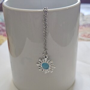 May include: A white ceramic mug with a silver chain and a silver sun charm with a blue center. The charm is hanging from the rim of the mug.