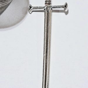 May include: A silver-toned metal sword charm attached to a fine link chain, featuring a detailed hilt and a straight blade. The charm is connected to a spherical stainless steel mesh tea infuser, which is partially visible in the upper left corner.
