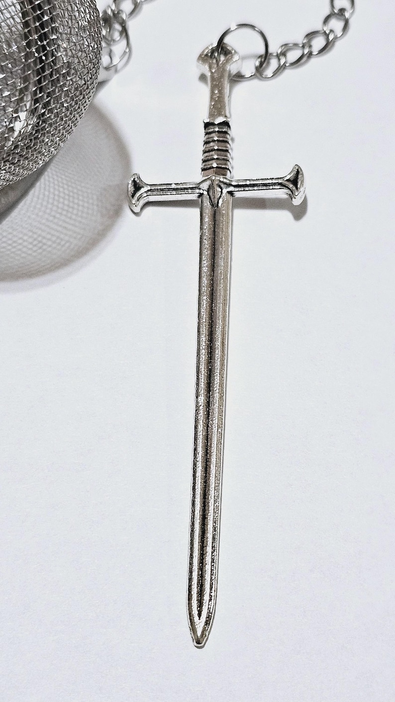 May include: A silver-toned metal sword charm attached to a fine link chain, featuring a detailed hilt and a straight blade. The charm is connected to a spherical stainless steel mesh tea infuser, which is partially visible in the upper left corner.