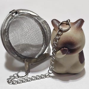May include: A silver tea infuser with a mesh ball and a chain attached to a ceramic cat figurine. The cat is beige with brown markings and has its paws clasped together.