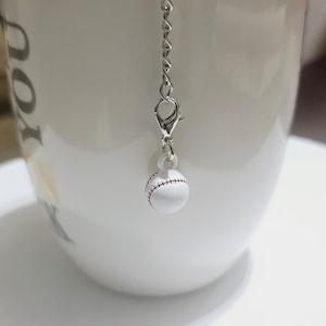 May include: A white baseball charm with red stitching hangs from a silver chain. The charm is attached to a white ceramic mug.