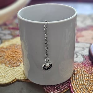 May include: A white ceramic mug with a silver chain and a decorative seashell-shaped tea infuser. The infuser has a black and silver design with a small white pearl in the center. The mug is sitting on a decorative beaded coaster.