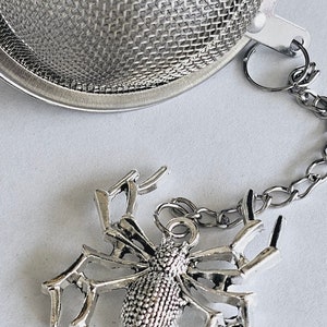 May include: A silver tea infuser shaped like a spider with a chain attached. The infuser is on a white surface.