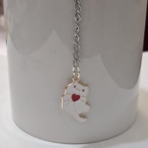 Puede incluir: Una taza de cerámica blanca con un infusor de té de cadena plateada. El infusor presenta un dije de gato blanco con un corazón rojo y ribete dorado. La taza es gris claro.