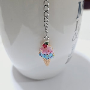 Può includere: Un infusore per tè a catena argentata con un ciondolo a forma di cono gelato in ceramica. Il cono gelato è decorato con palline di gelato rosa e blu con codette e una ciliegia rossa in cima.