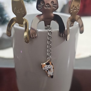 May include: Three silver cat-shaped tea infusers with a dangling charm of a cat. The infusers are hanging on the rim of a white mug.