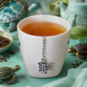 Peut inclure: Une tasse en céramique blanche remplie de thé ambré, avec une chaîne d'infuseur à thé en métal argenté suspendue sur le bord. La breloque de l'infuseur a la forme d'une petite tortue avec des cristaux clairs incrustés sur sa carapace.