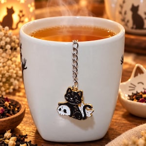 Peut inclure: Une tasse en céramique blanche remplie de thé fumant, avec une chaîne d'infuseur à thé en métal attachée à une breloque en émail noir et blanc représentant un chat assis à côté d'un crâne. La breloque a des bords dorés et des ailes blanches.